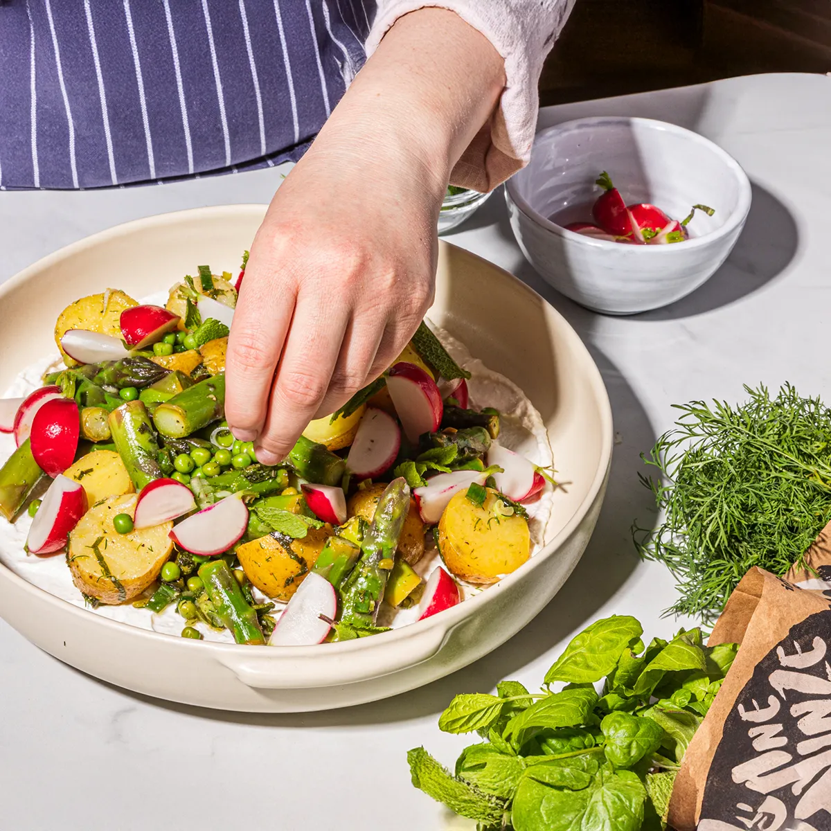 Hand placing peas on a fresh vegetable salad with potatoes, radishes, and greens in a white bowl.