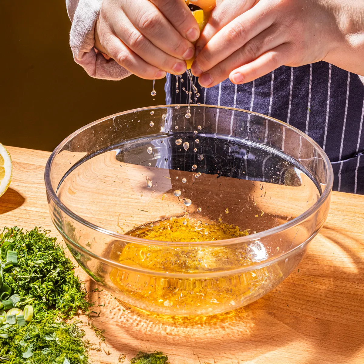 Hands squeezing lemon juice into a glass bowl with chopped herbs on a wooden cutting board.
