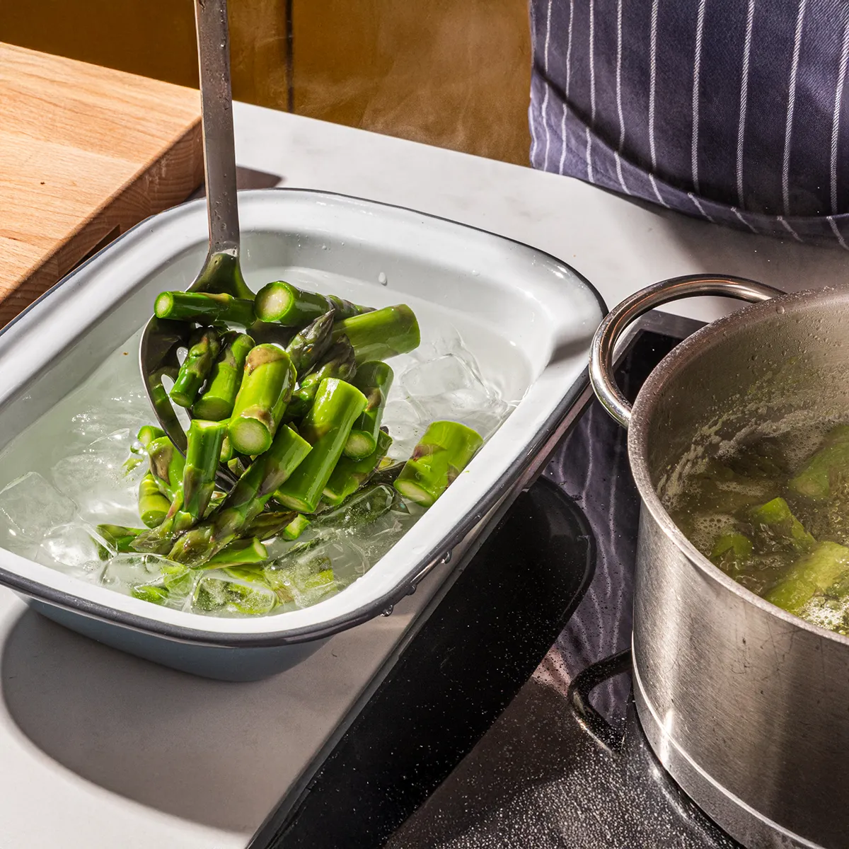 Cooked asparagus being transferred from boiling water to an ice bath for blanching, with a pot and stovetop visible.