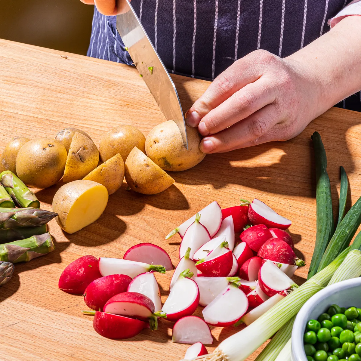 Hands slicing potatoes on a board surrounded by chopped radishes, asparagus, green onions, and a bowl of green peas.