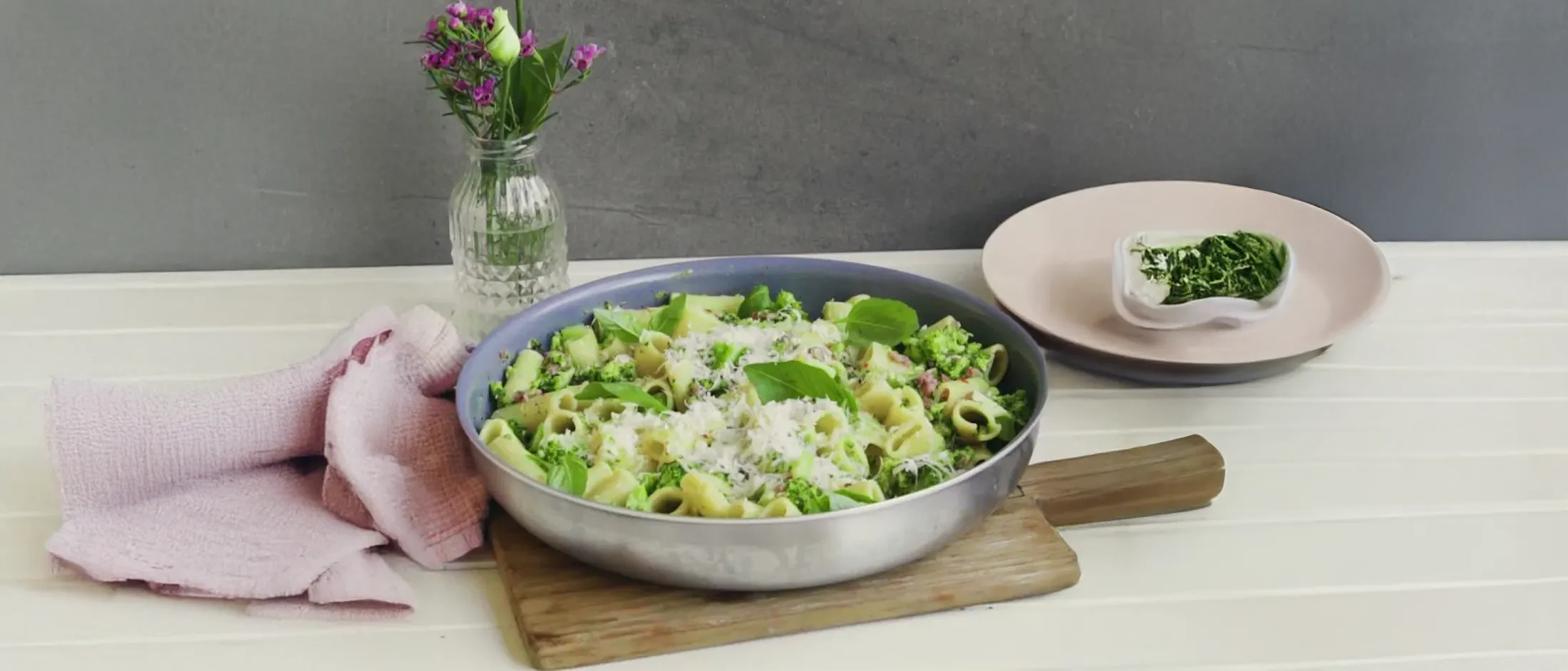 Hands grating a block of hard cheese over a pan of rigatoni pasta with broccoli and bits of meat on a stovetop.