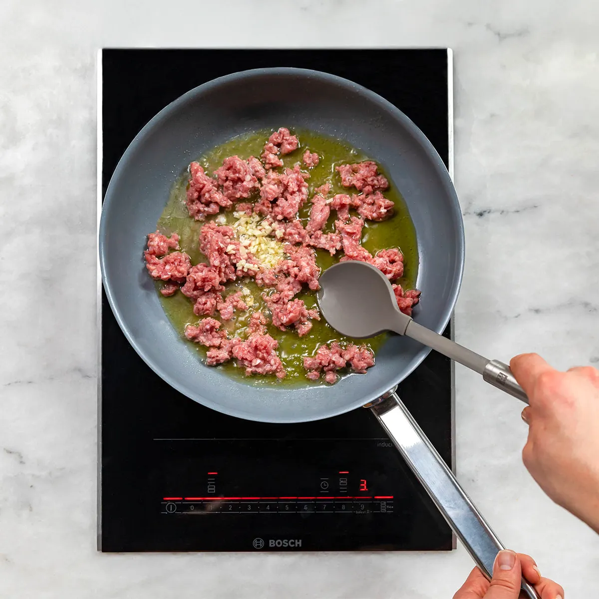 Sausages cooking in a frying pan with oil and minced garlic on an induction stove with a hand stirring with a spoon.