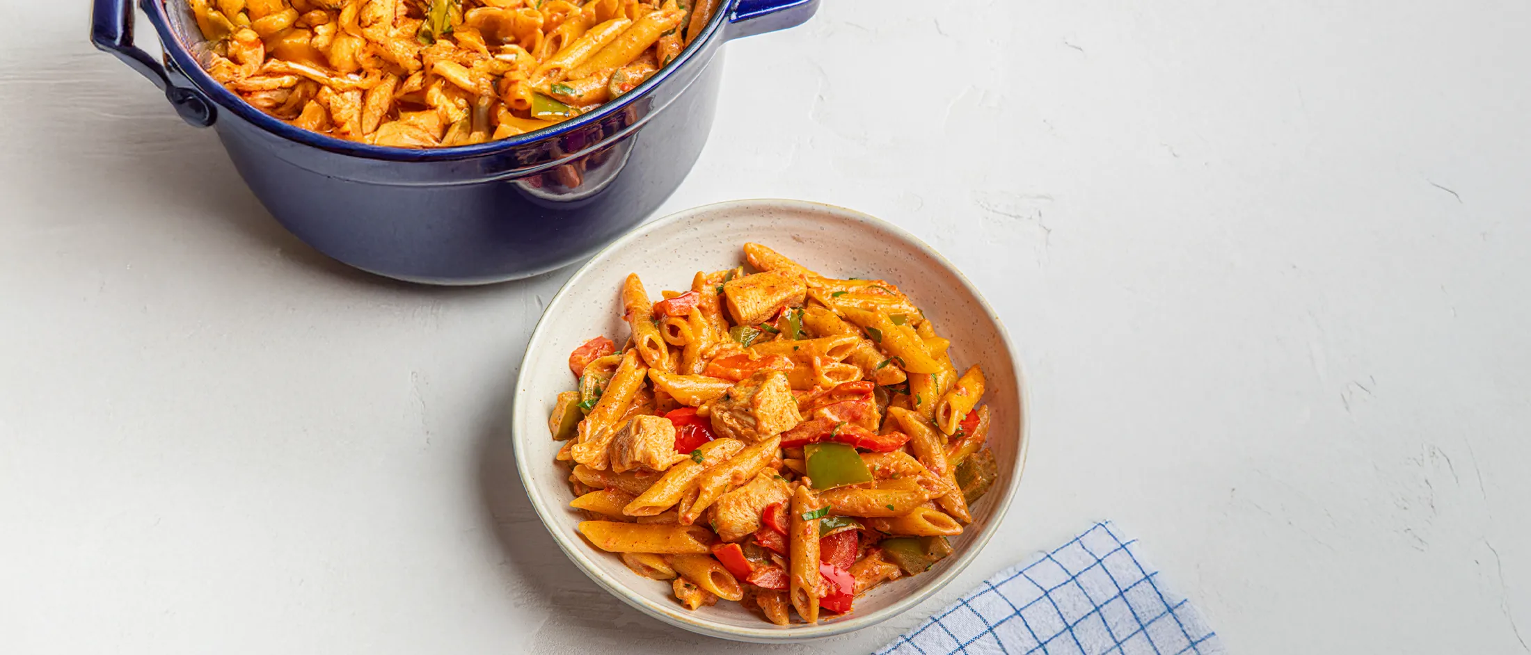 Bowl of creamy penne pasta with chicken, red and green bell peppers, garnished with herbs, next to a blue pot on a white surface.