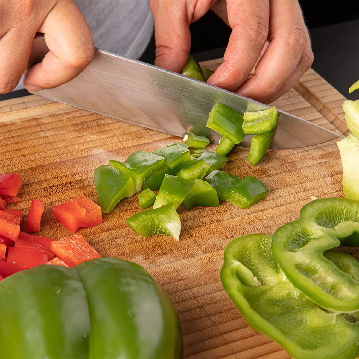 Hands chopping green bell pepper on a wooden cutting board with sliced red and green bell peppers nearby.