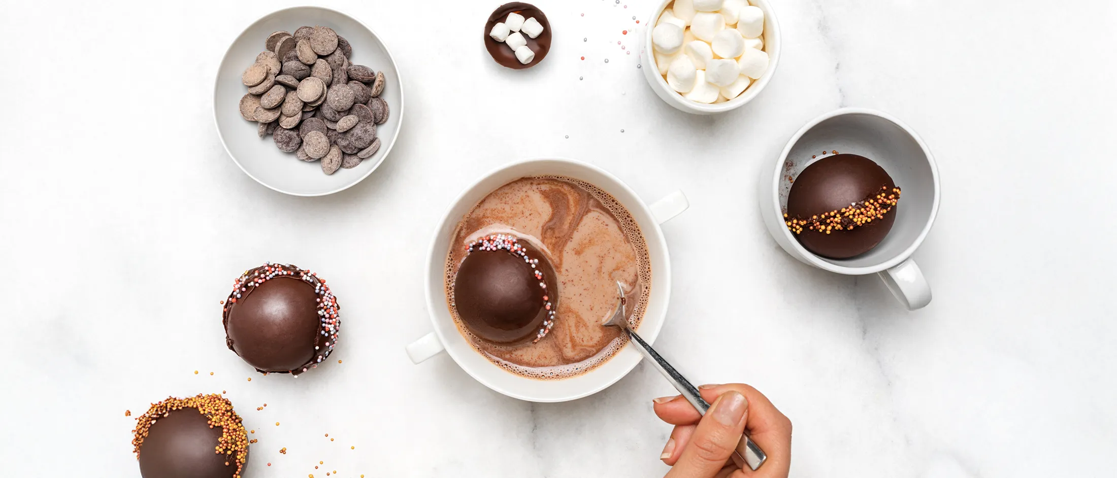 Overhead view of a hand stirring a mug of hot chocolate as a chocolate cocoa bomb melts inside, surrounded by bowls of chocolate wafers, mini marshmallows, and sprinkle-topped cocoa bombs on a white marble surface.
