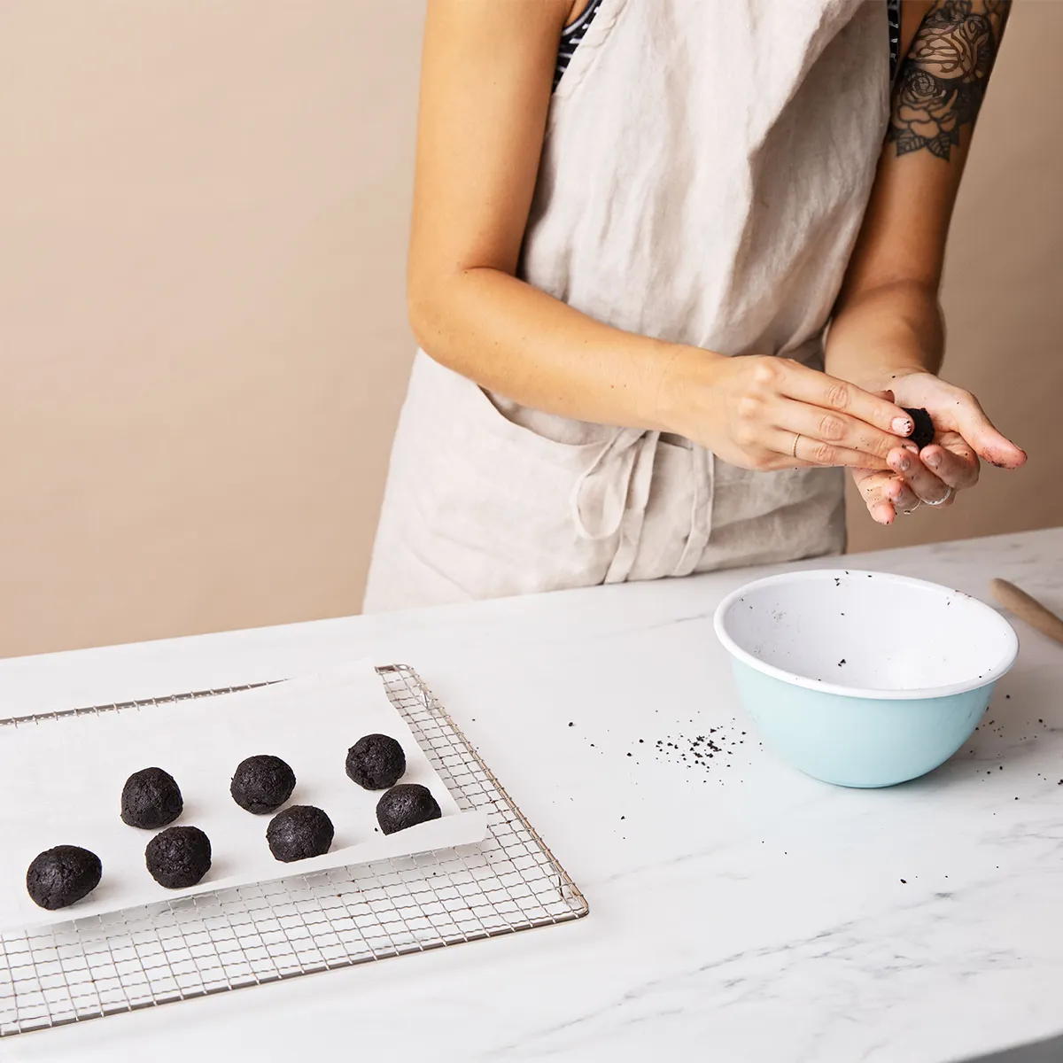 Person shaping black dough balls at a white counter with bowls and a cooling rack, wearing a beige apron