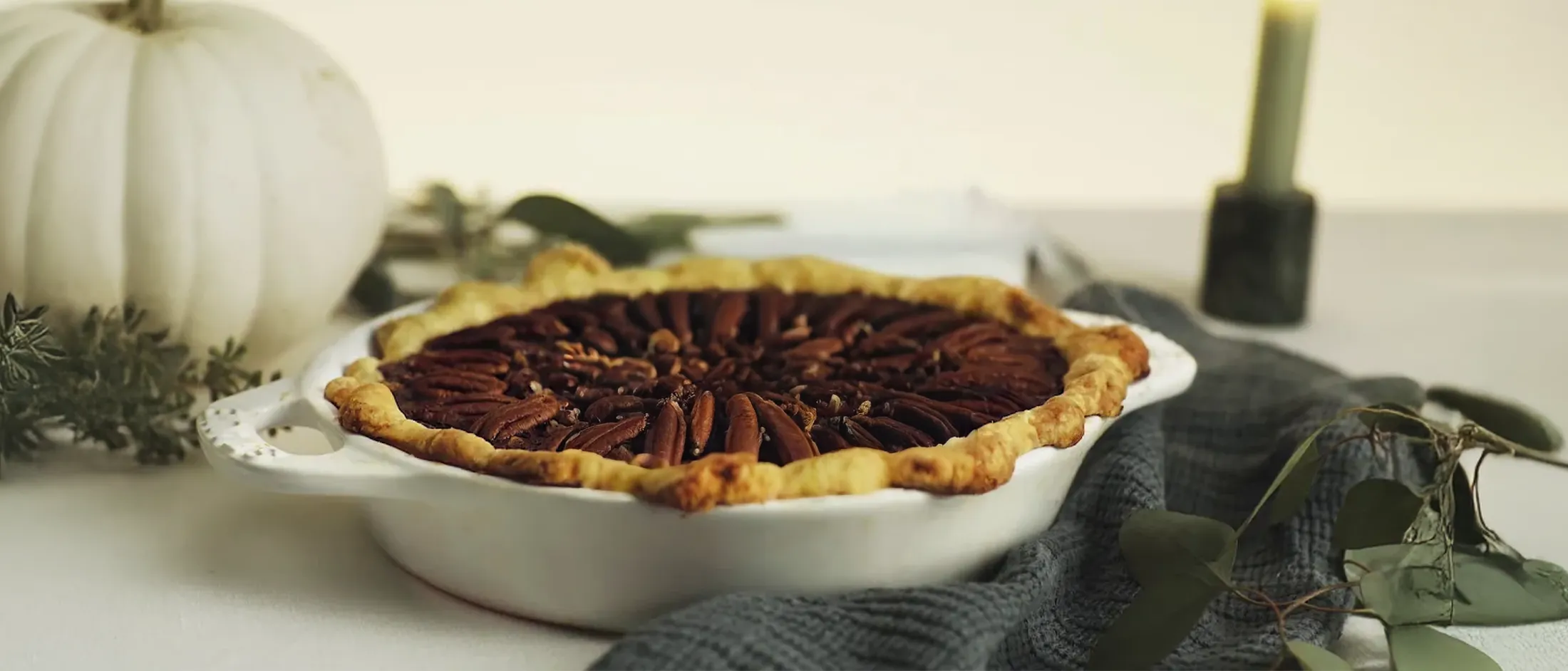 Freshly baked pecan pie in a white ceramic pie dish on a soft blue cloth with a white pumpkin in the background