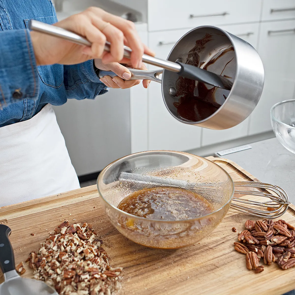 Hands pouring melted chocolate into caramel in a glass bowl, with chopped and whole pecans on a wooden cutting board