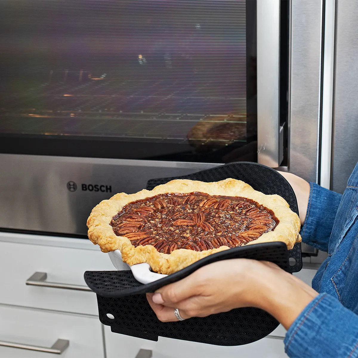 Hands in blue sleeves with oven mitts hold a freshly baked pecan pie in front of a stainless-steel Bosch oven