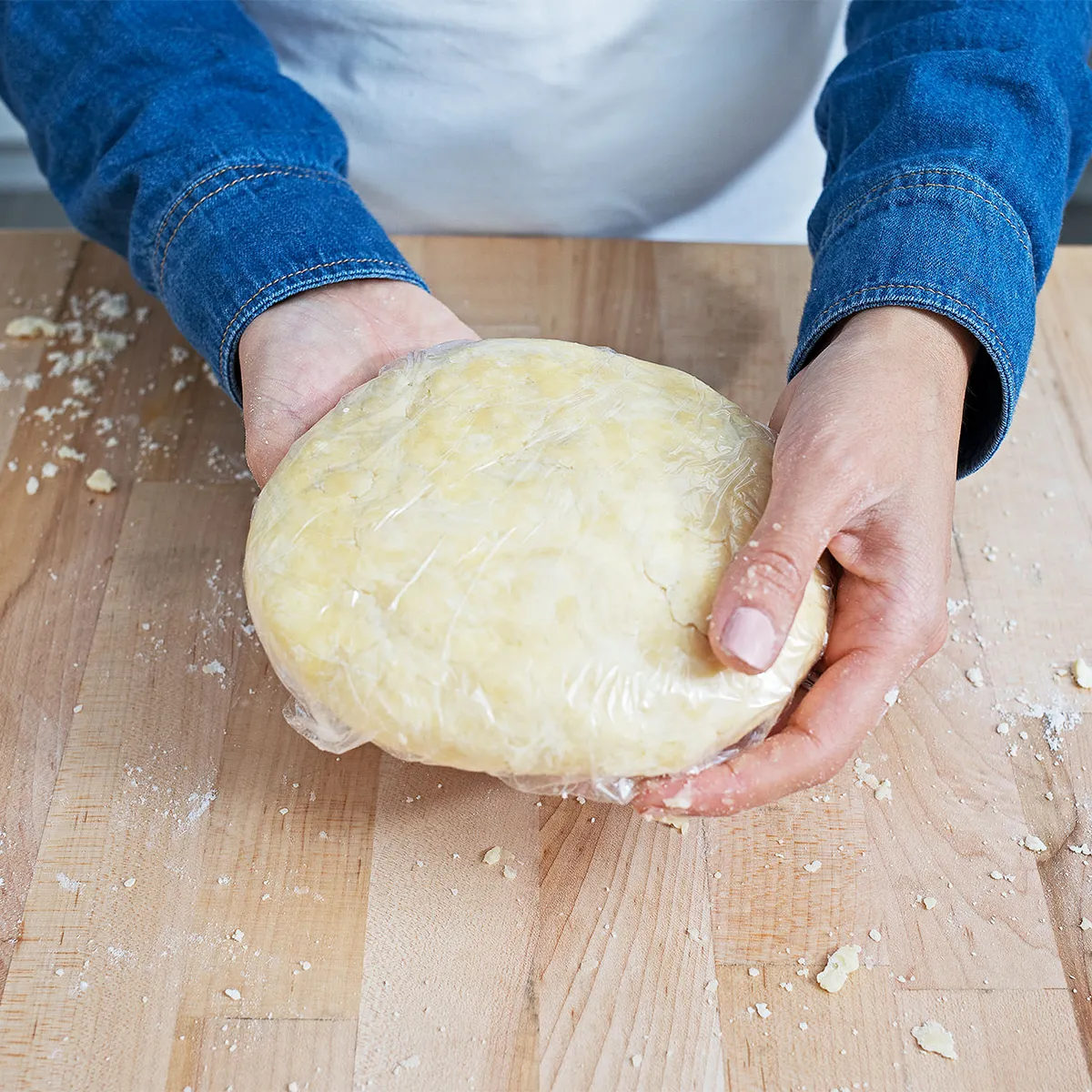 Hands holding a plastic-wrapped pie dough disk on a floured wooden board