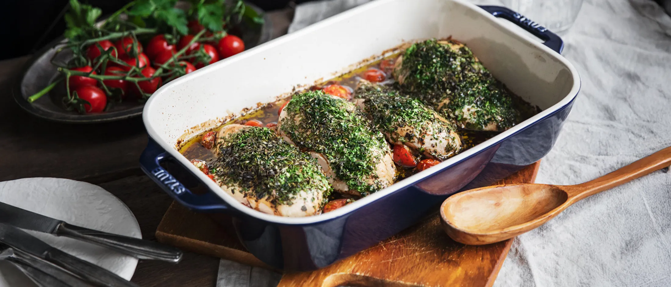 Baked herb-crusted chicken breasts with cherry tomatoes in a blue and white ceramic baking dish on a wooden board.