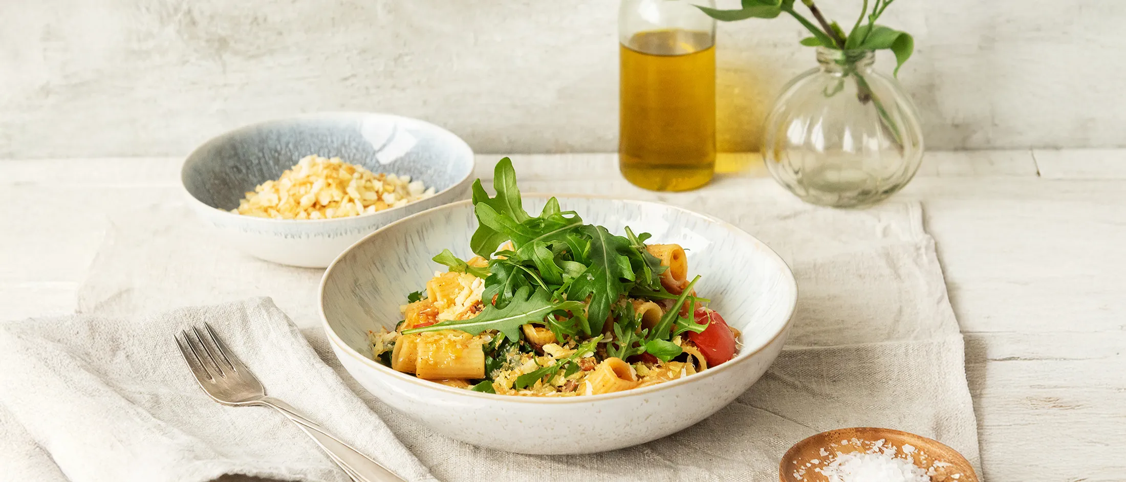 Bowl of pasta with arugula on top on a tablecloth next to a fork on a napkin, a bowl of grated cheese, a bottle of oil, and a small dish of salt.