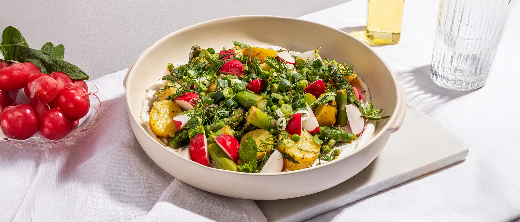Bowl of fresh salad with potatoes, radishes, asparagus, and herbs on a white tablecloth with olive oil bottle nearby.