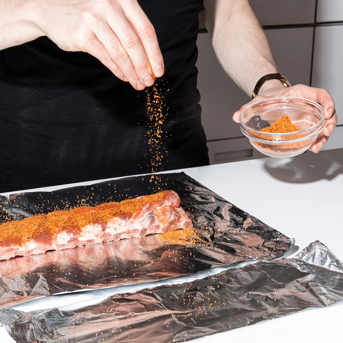 A person in a black shirt sprinkles a reddish-orange spice rub from their hand onto a rack of ribs resting on aluminum foil, with a small bowl of the rub nearby.