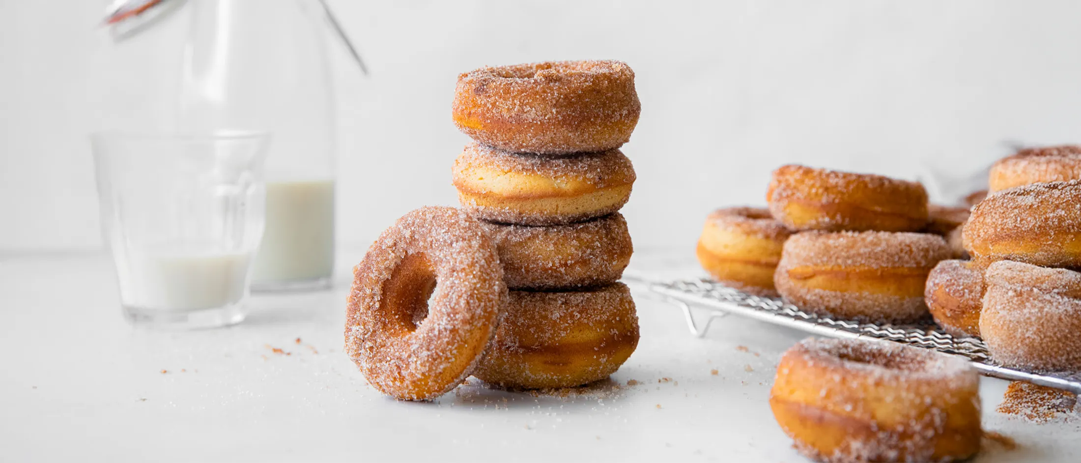 Stack of sugar-coated cinnamon donuts with a milk bottle and glass in the background on a white surface.
