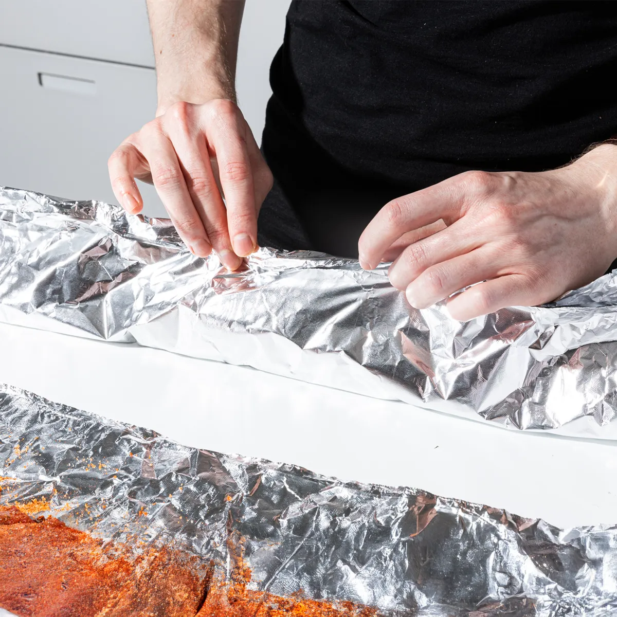 Person sprinkling seasoning onto raw ribs placed on foil on a kitchen counter, holding a small bowl of spice blend.