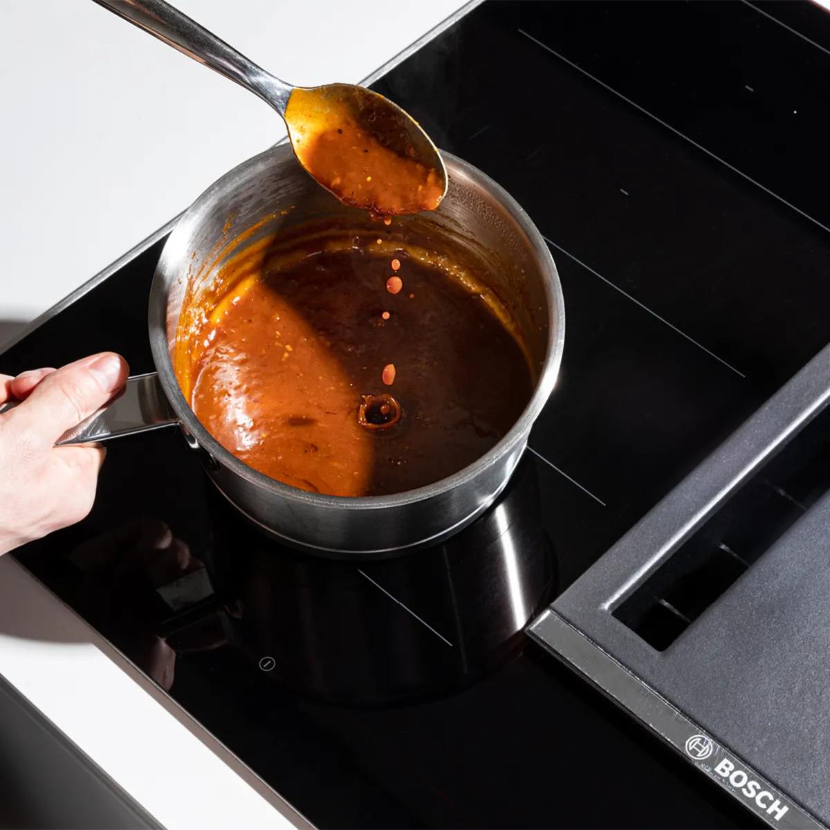 A hand holds a spoon, dripping rich, reddish-brown sauce back into a small saucepan simmering on a sleek black Bosch induction cooktop.