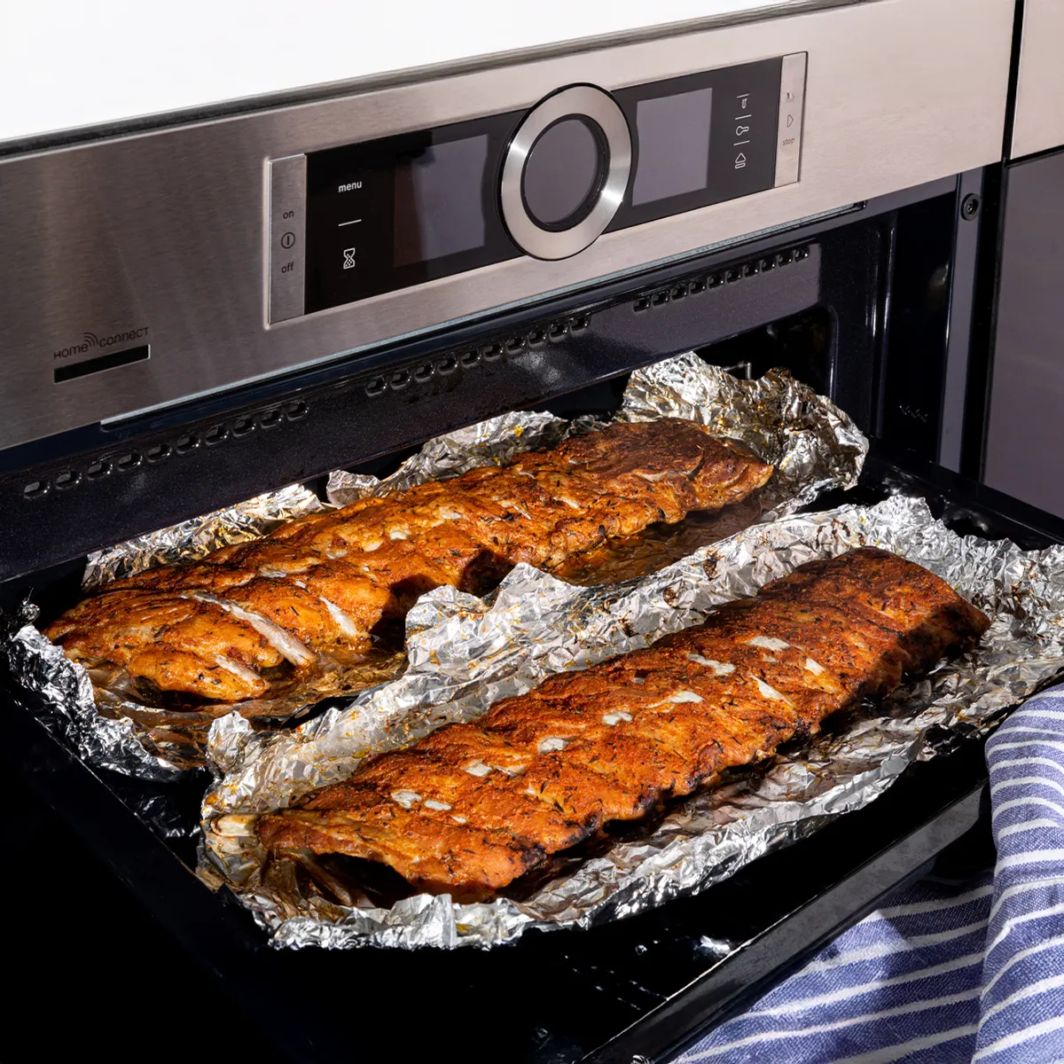 Two racks of seasoned ribs, wrapped in aluminum foil, are baking inside a modern stainless steel oven with a digital display.
