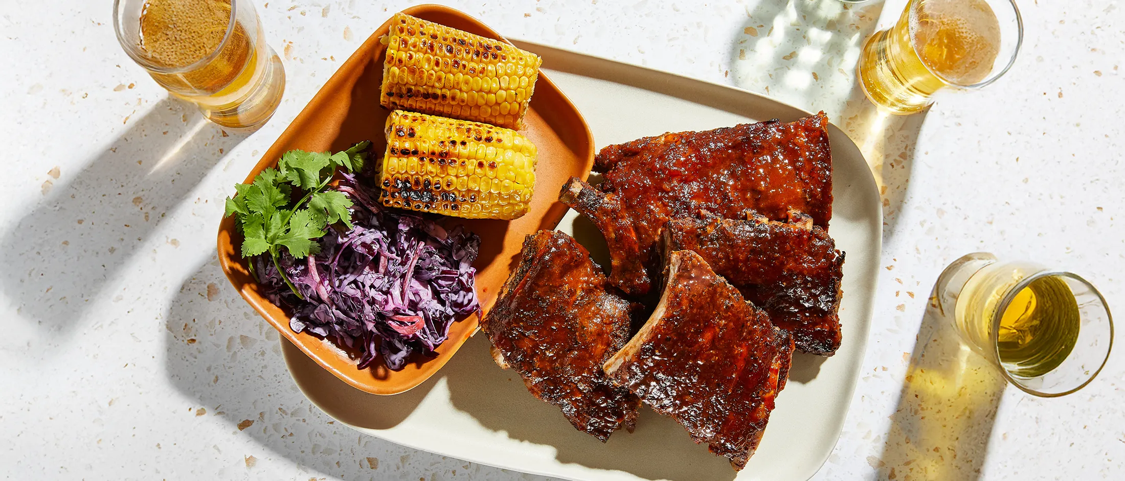 A delicious meal featuring grilled ribs slathered in BBQ sauce, two charred corn on the cob, and a vibrant purple cabbage slaw with cilantro.