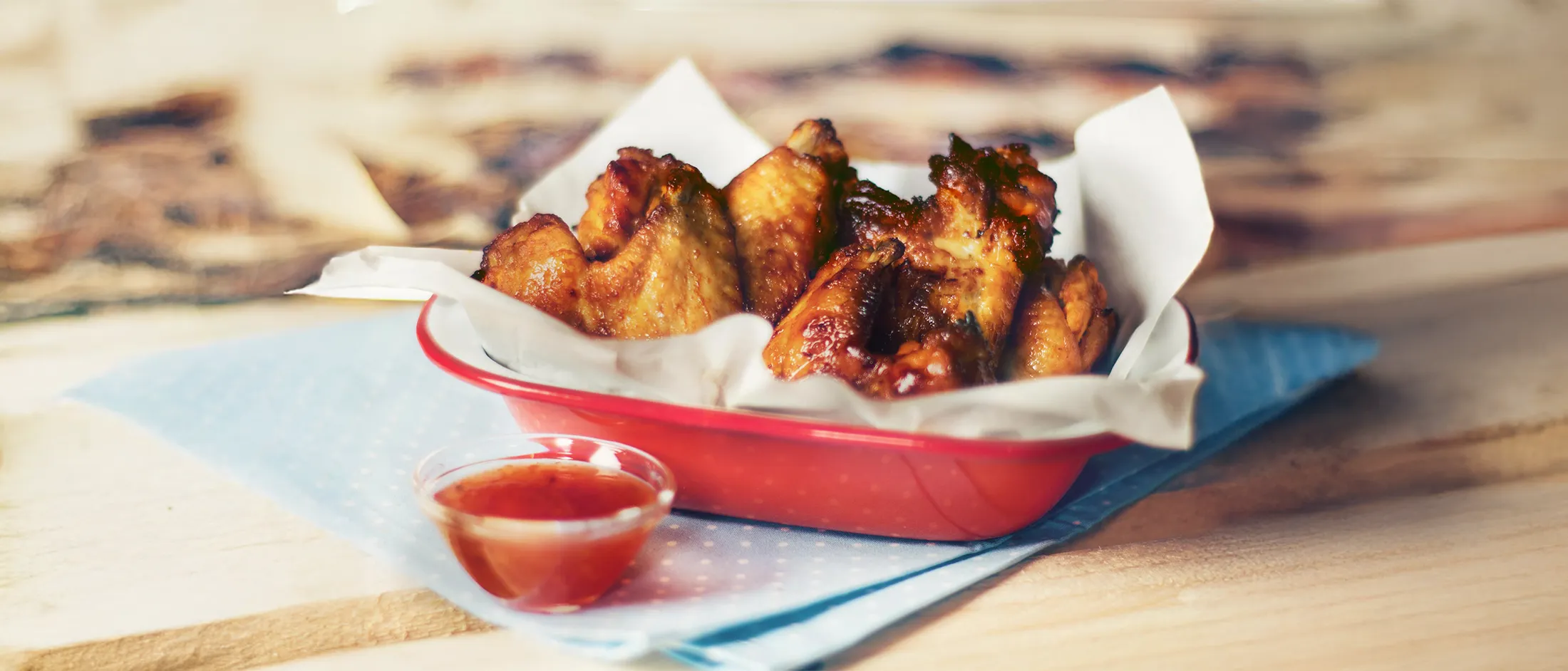Crispy chicken wings in a red tray lined with parchment paper, accompanied by a small cup of red dipping sauce on a light wooden table.
