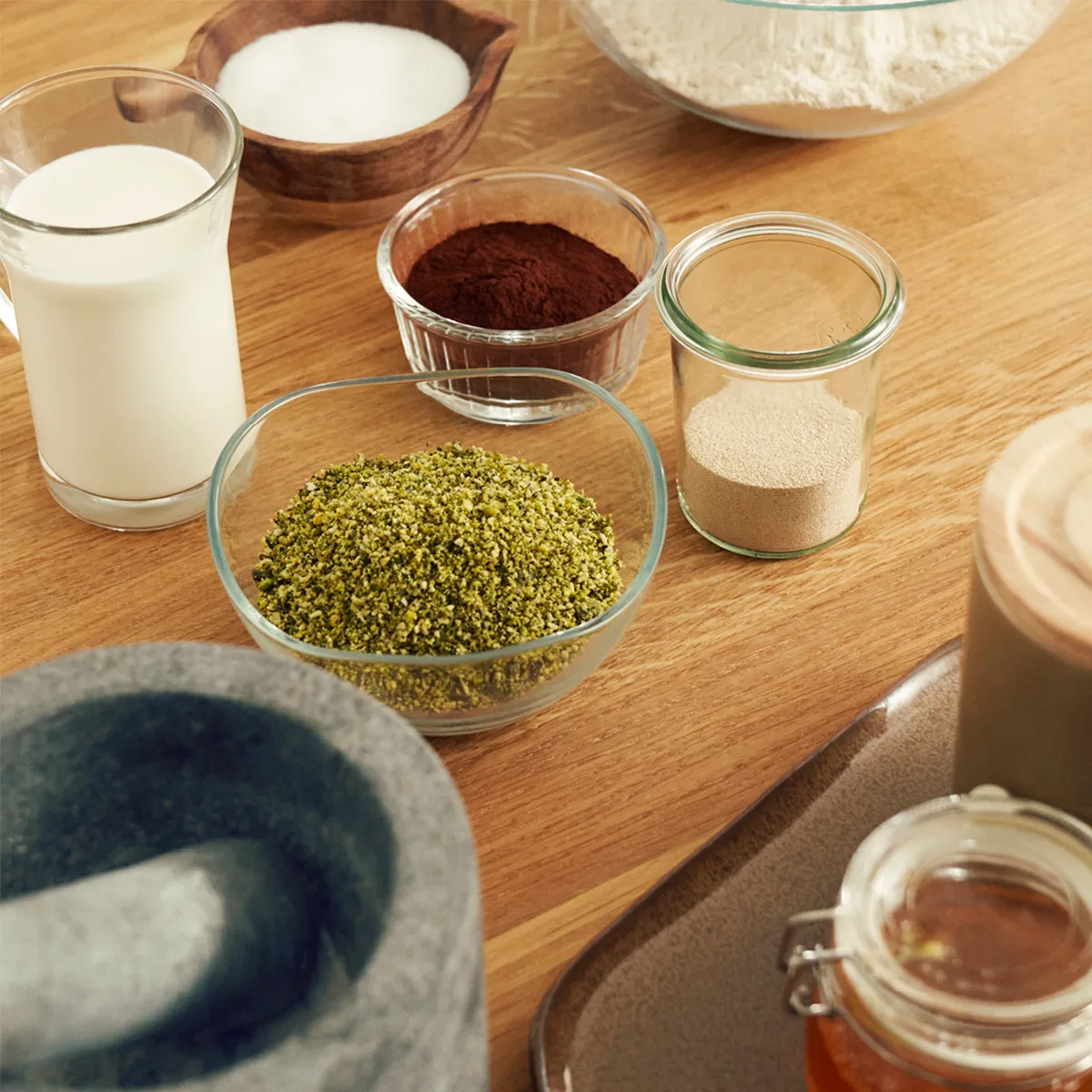 Various baking ingredients in glass bowls and jars on a wooden surface, including milk, cocoa powder, yeast, and green herbs.