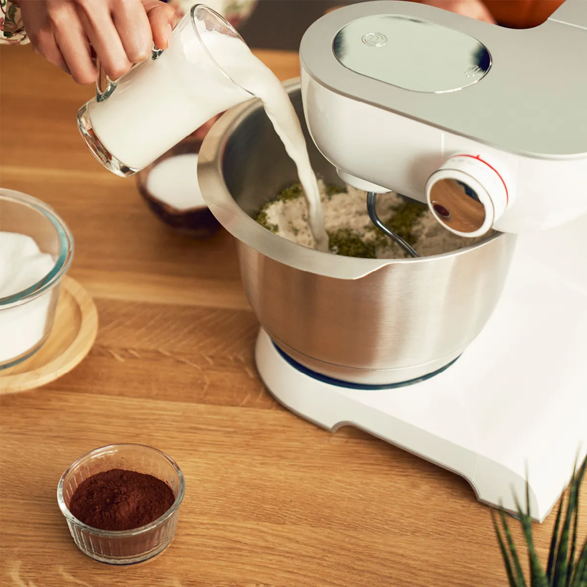Hand pouring milk into a stand mixer bowl with dry ingredients on a wooden countertop, alongside cocoa powder and sugar bowls.