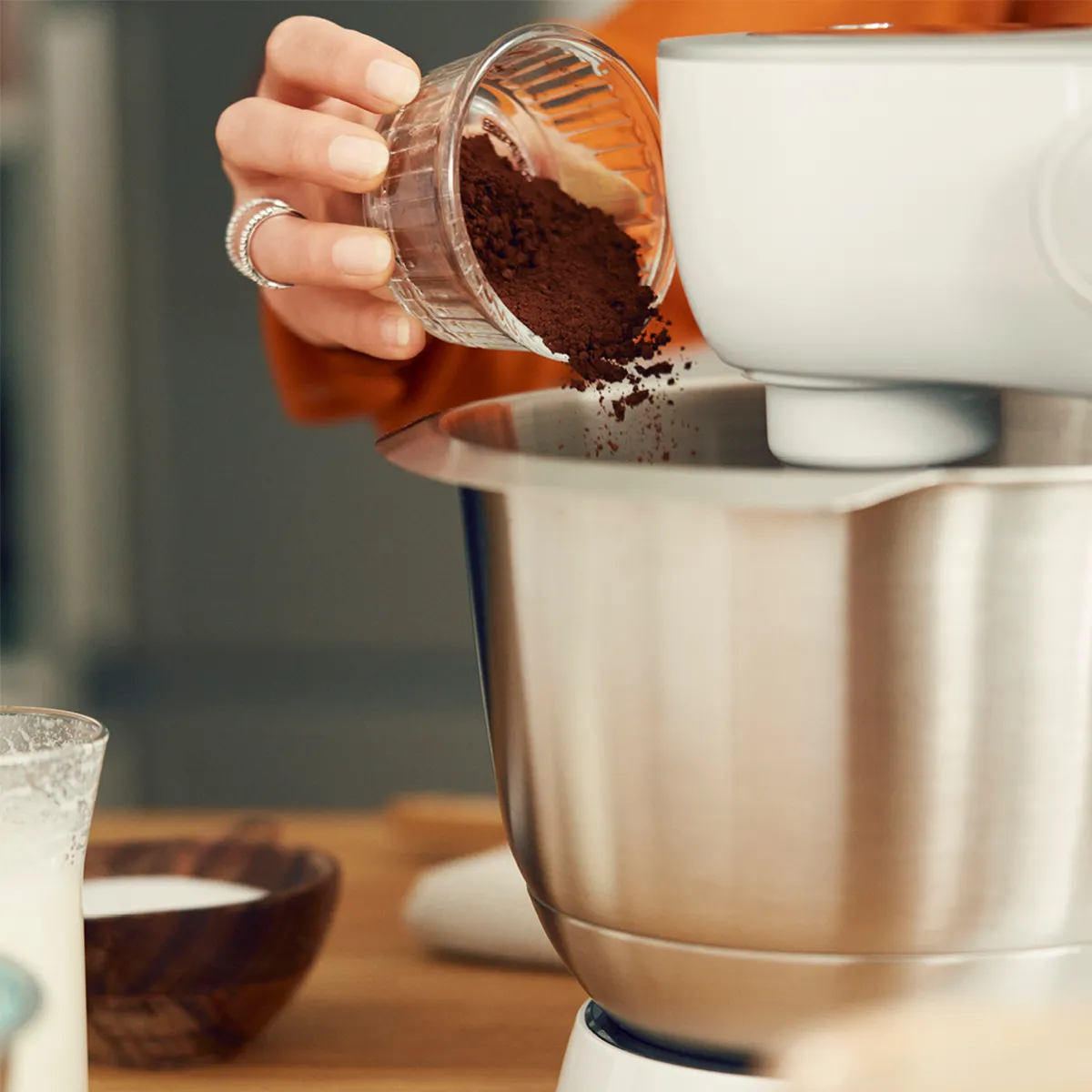 Hand pouring  cocoa powder from a glass container into  into a stand mixer bowl.