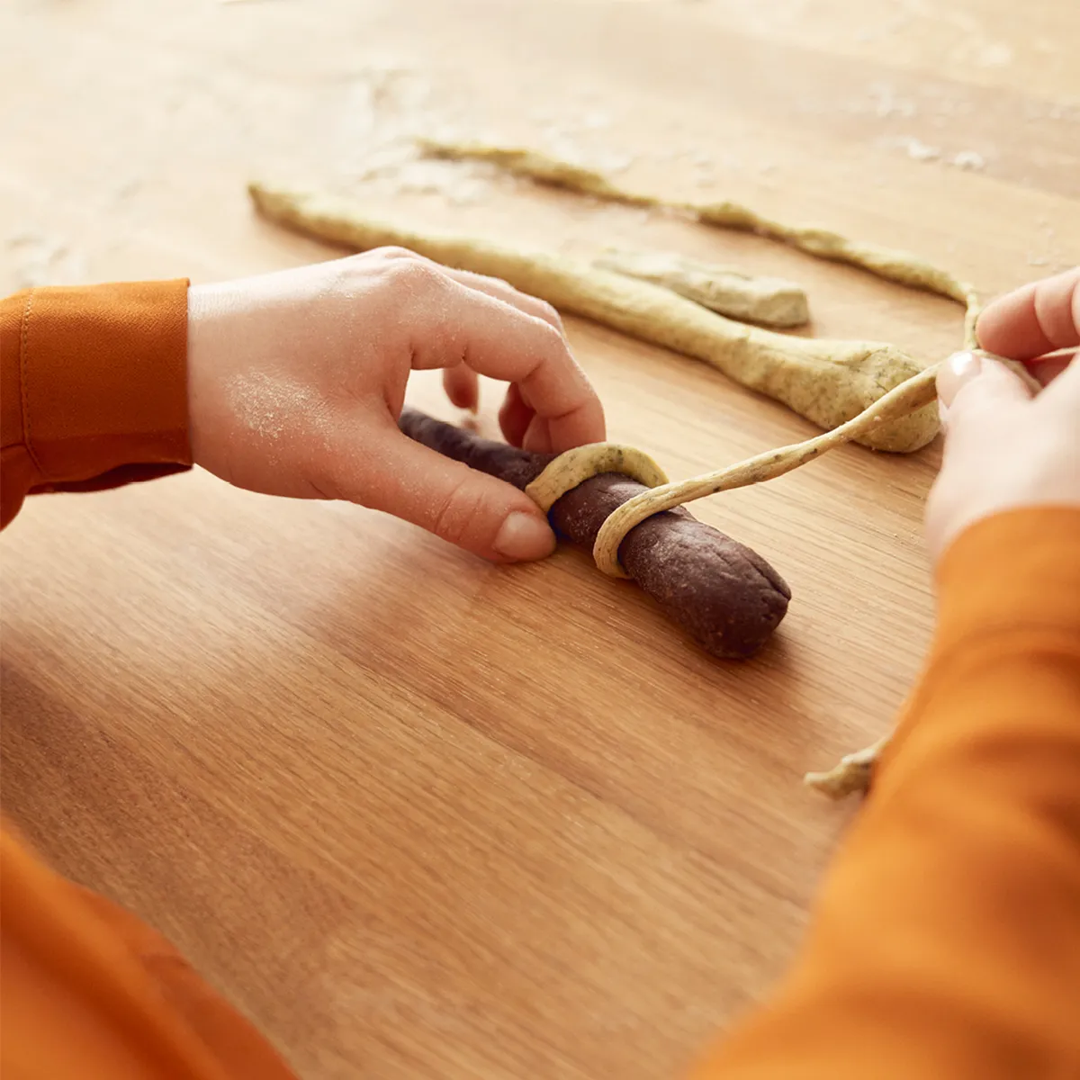 Hands wrapping a strip of dough around a darker dough on a wooden surface, preparing chocolate pretzel sticks.
