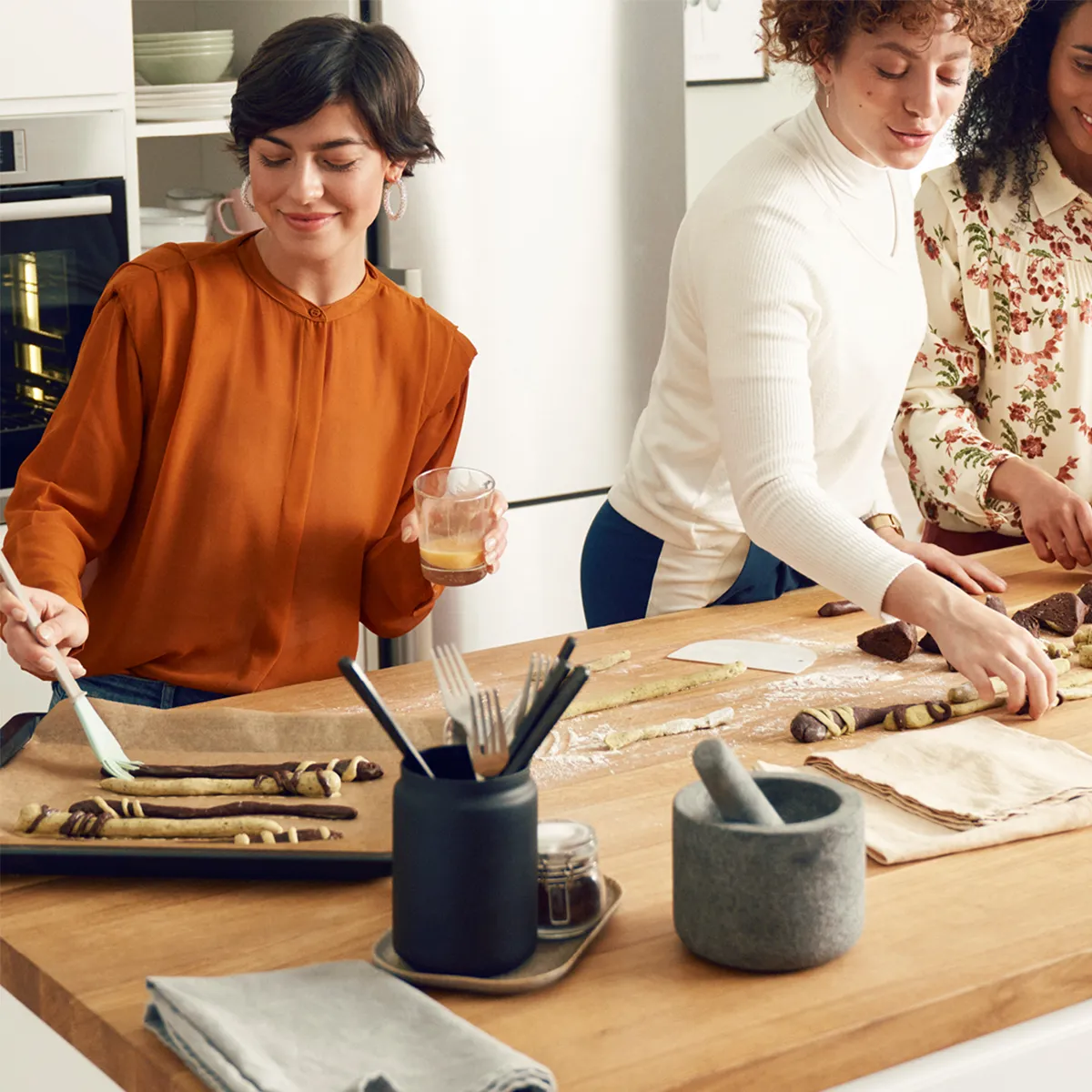 Three people preparing and decorating chocolate pretzel sticks on a kitchen counter with baking tools and ingredients.