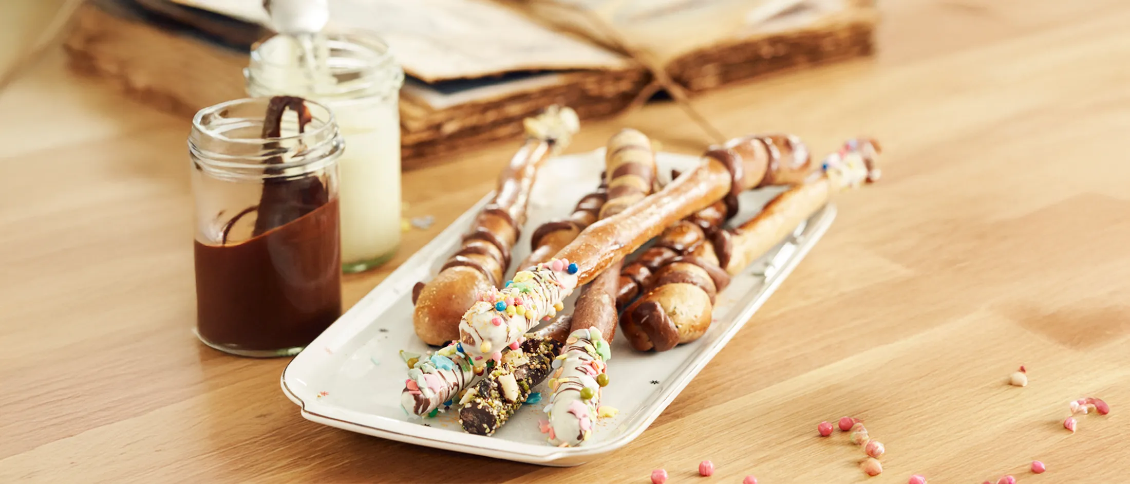 Plate of assorted chocolate-dipped pretzel sticks decorated with sprinkles and nuts on a wooden table with open cookbooks.