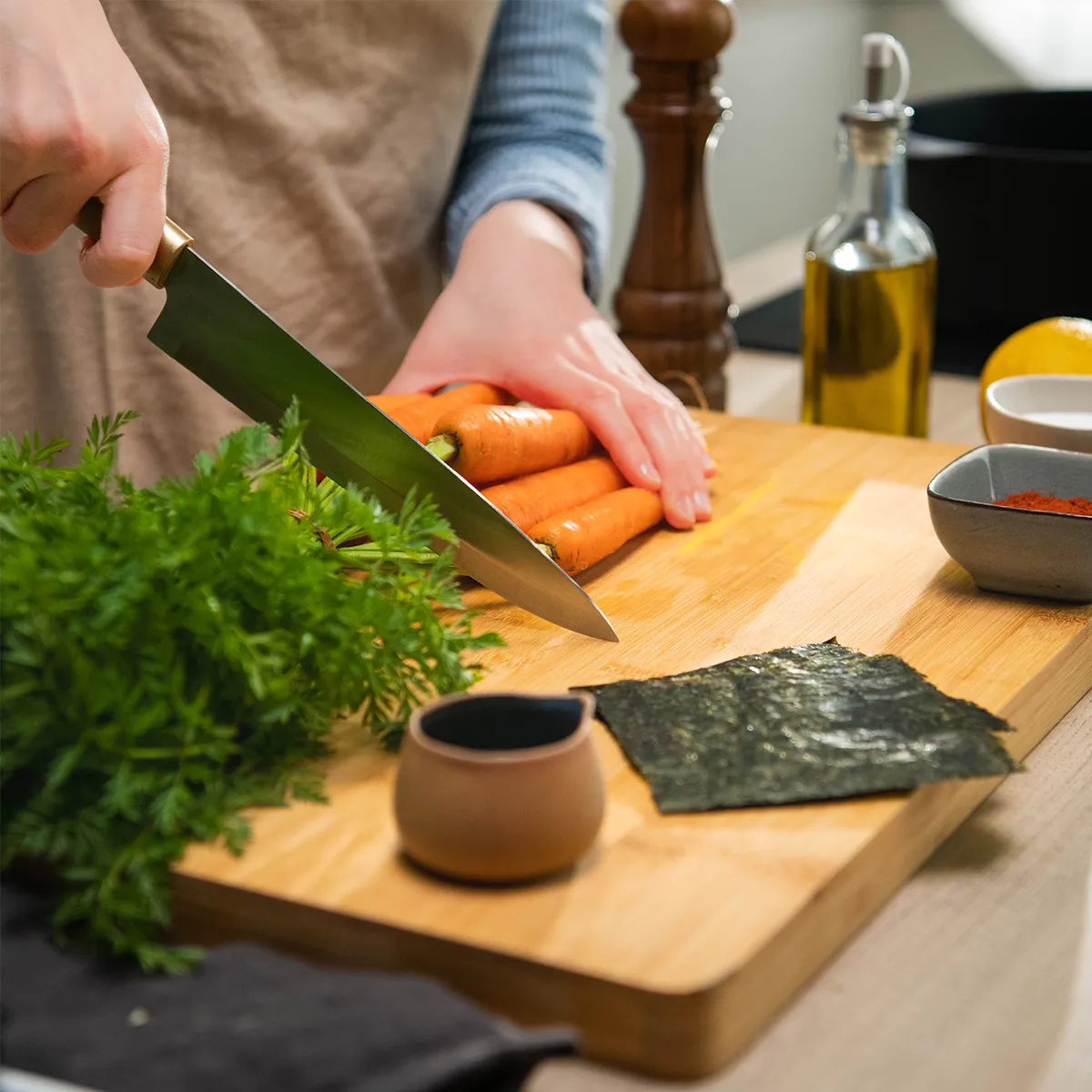Hands slicing a bunch of carrots on a wooden cutting board with a chef’s knife; carrot greens, a sheet of nori, small bowls of spices, a pepper mill and a bottle of olive oil sit nearby.