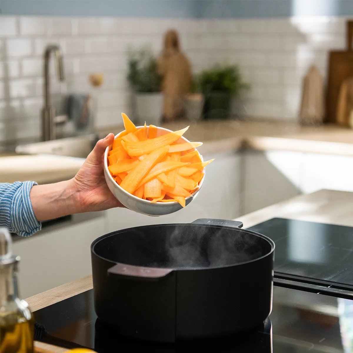Hands slicing a bunch of carrots on a wooden cutting board with a chef’s knife; carrot greens, a sheet of nori, small bowls of spices, a pepper mill and a bottle of olive oil sit nearby.