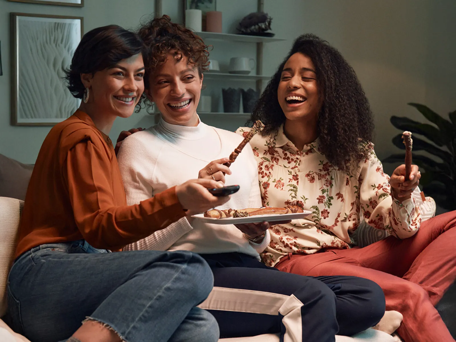 Three women sitting on a couch eating pretzel sticks in a cozy living room setting.