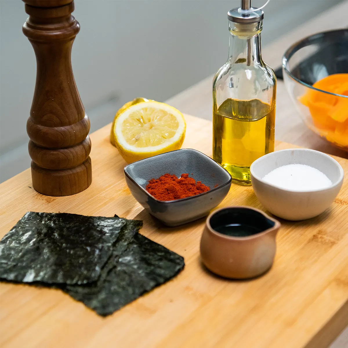 Wooden cutting board with a pepper mill, half a lemon, a bottle of oil, small bowls of red spice and salt, a tiny jug of dark sauce, several sheets of nori seaweed, and a glass bowl with bright orange vegetable ribbons.