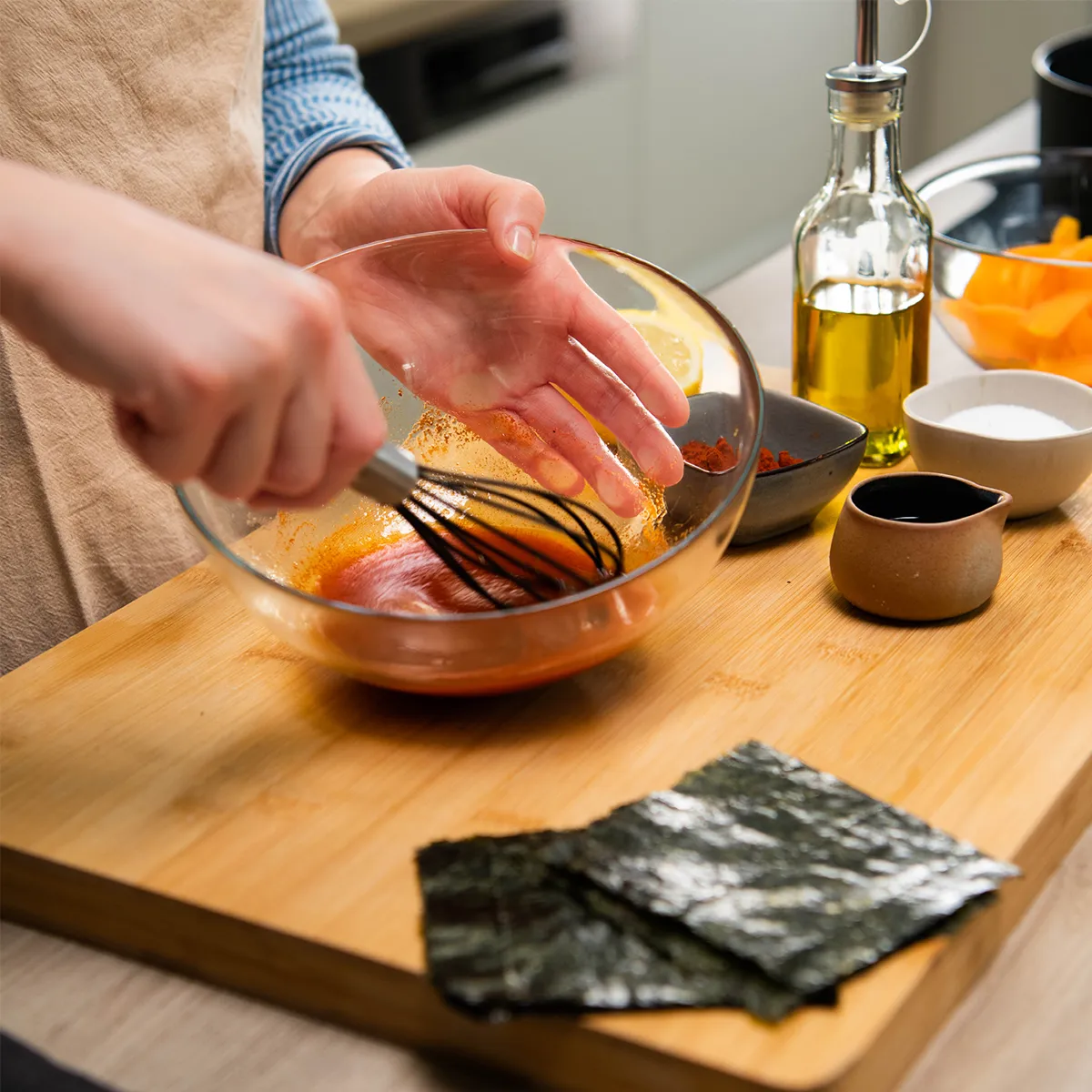 Person in an apron whisking a red-orange sauce in a glass bowl on a wooden board, with bowls of spices, oil, a small jug of dark sauce and seaweed sheets nearby