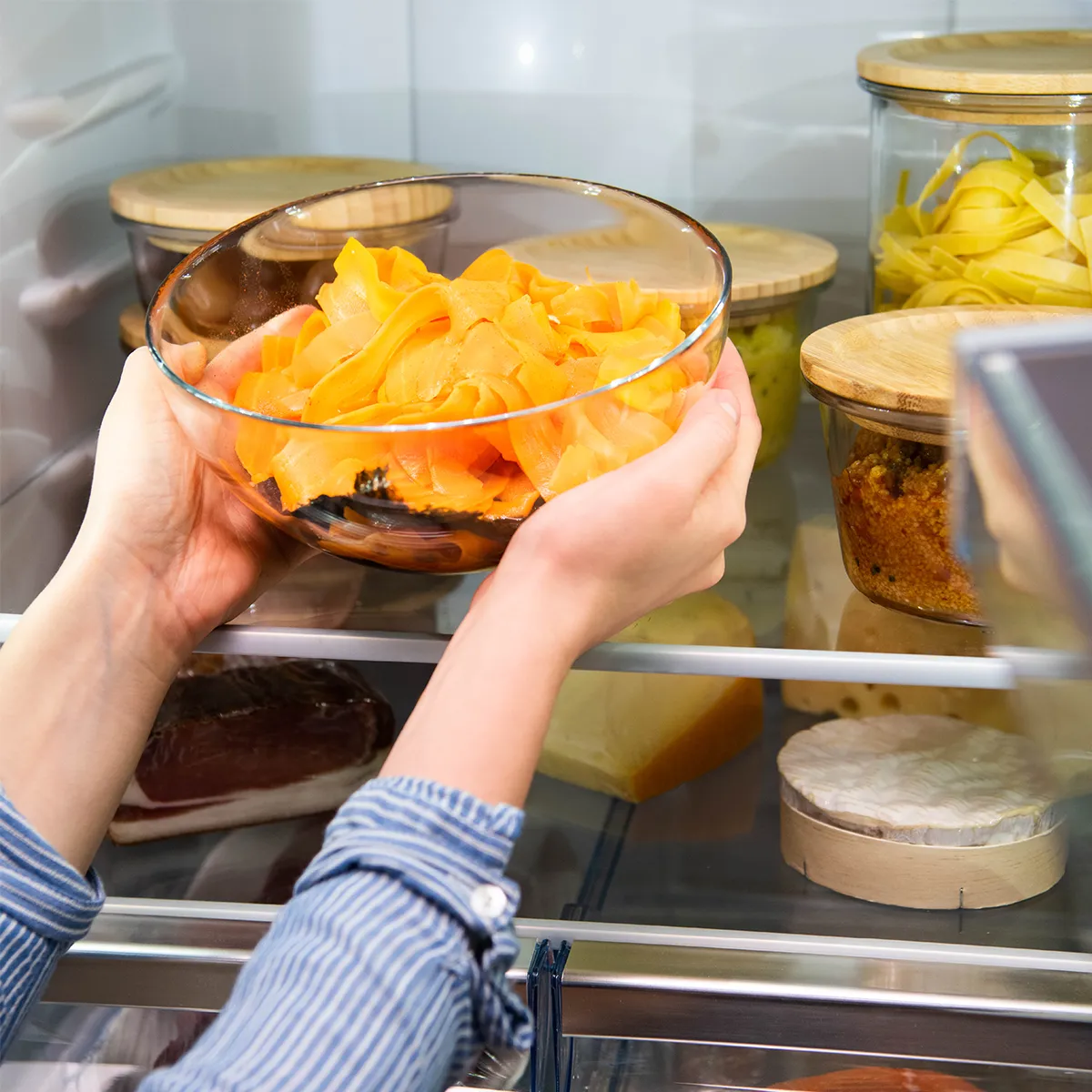 Hands setting a glass bowl of thin orange vegetable ribbons onto a refrigerator shelf, surrounded by jars with wooden lids and cheeses.