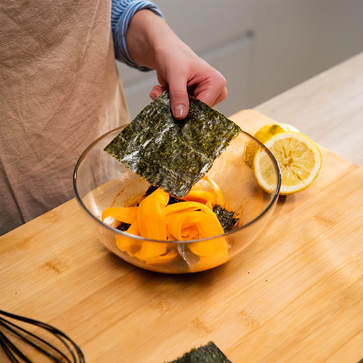 Close-up of hands placing a square sheet of seaweed into a glass bowl of orange vegetable ribbons, with a halved lemon visible on the counter.