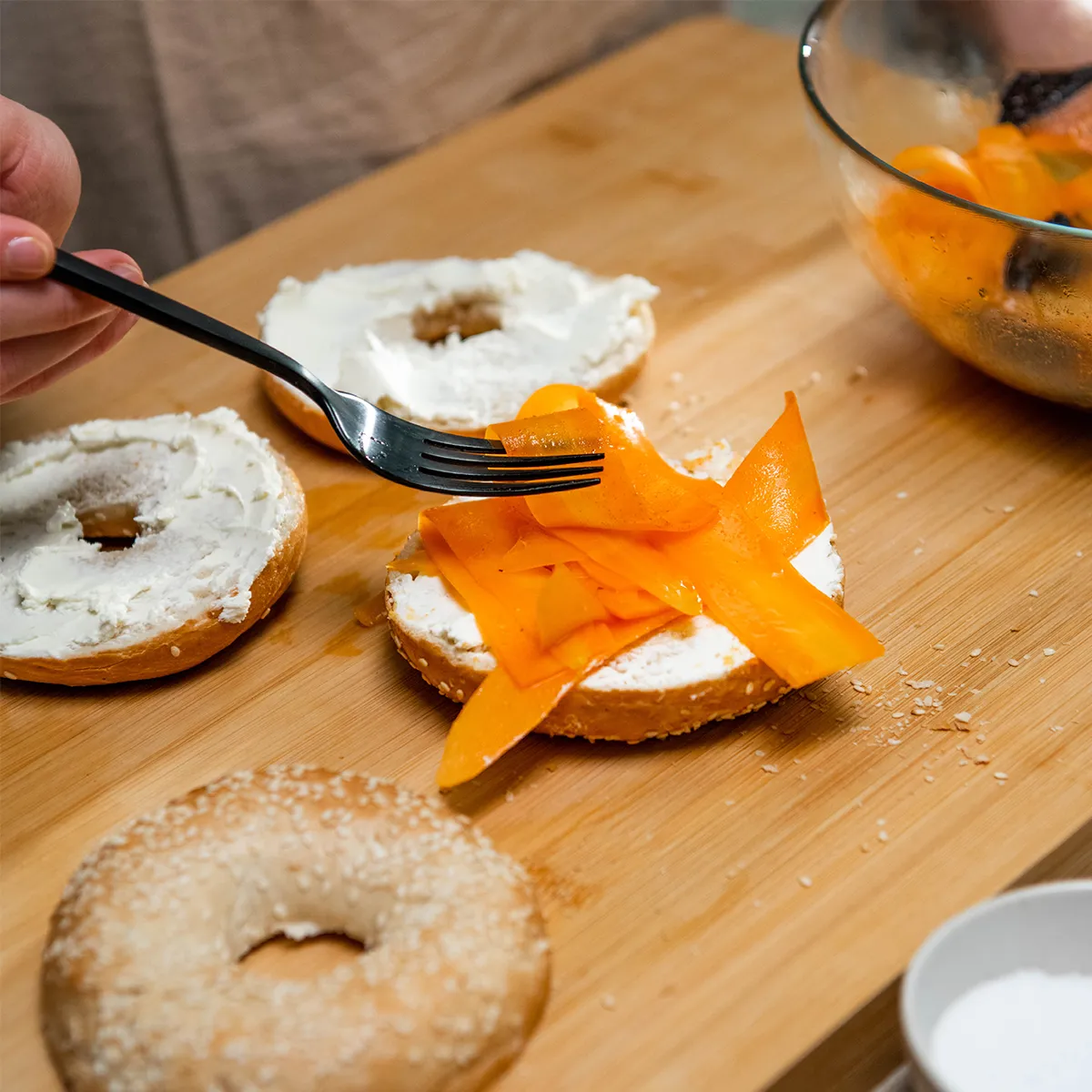 A fork arranging thin orange vegetable ribbons onto a cream cheese–spread bagel half on a wooden board, with another bagel half and the bowl of ribbons nearby.