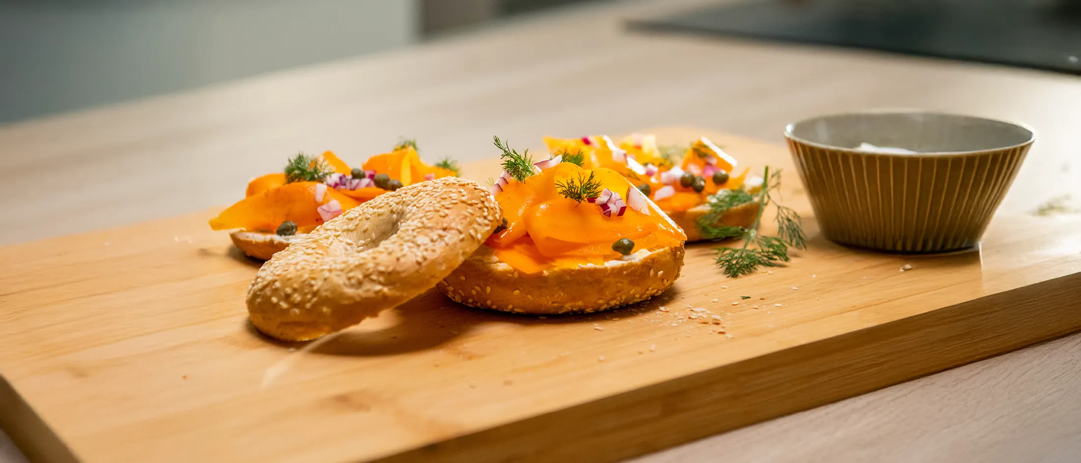 Sesame-seed bagel halves topped with folded smoked salmon, chopped red onion, capers and fresh dill on a wooden cutting board in a kitchen.
