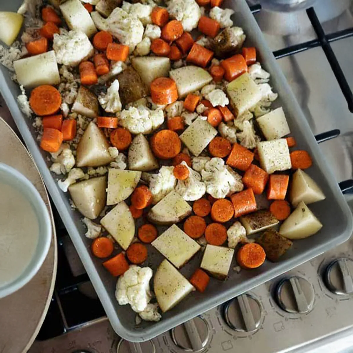 Sheet pan filled with seasoned chopped potatoes, carrots, and cauliflower ready for roasting on a stovetop.