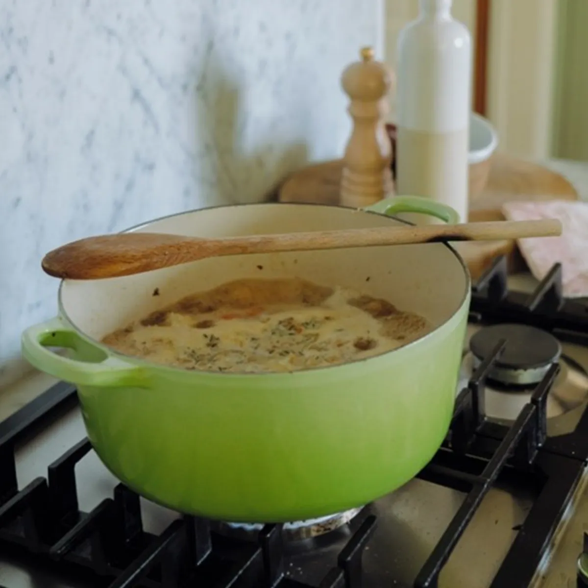 Green Dutch oven on a gas stove with a wooden spoon resting on top, simmering a creamy stew or sauce.