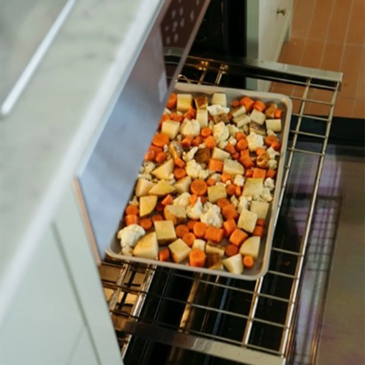 Tray of mixed chopped vegetables, including carrots, cauliflower, and potatoes, being placed into a modern stainless steel oven.