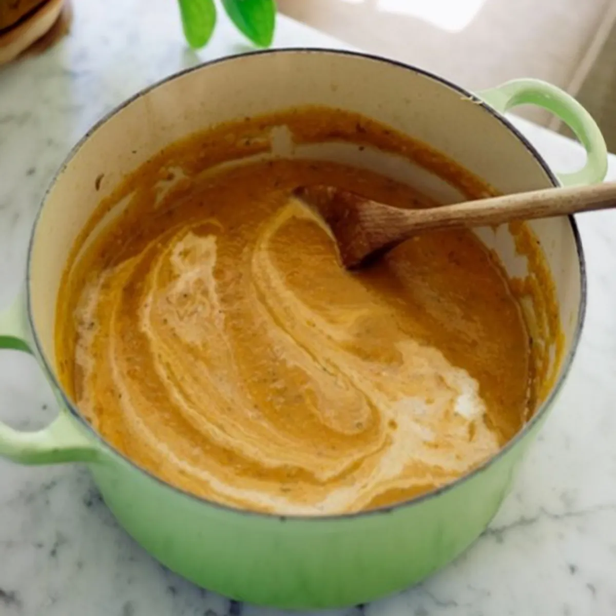 Green enameled pot of creamy orange soup swirled with cream and a wooden spoon, sitting on a marble countertop; potted plant and sunlit couch blurred in background.