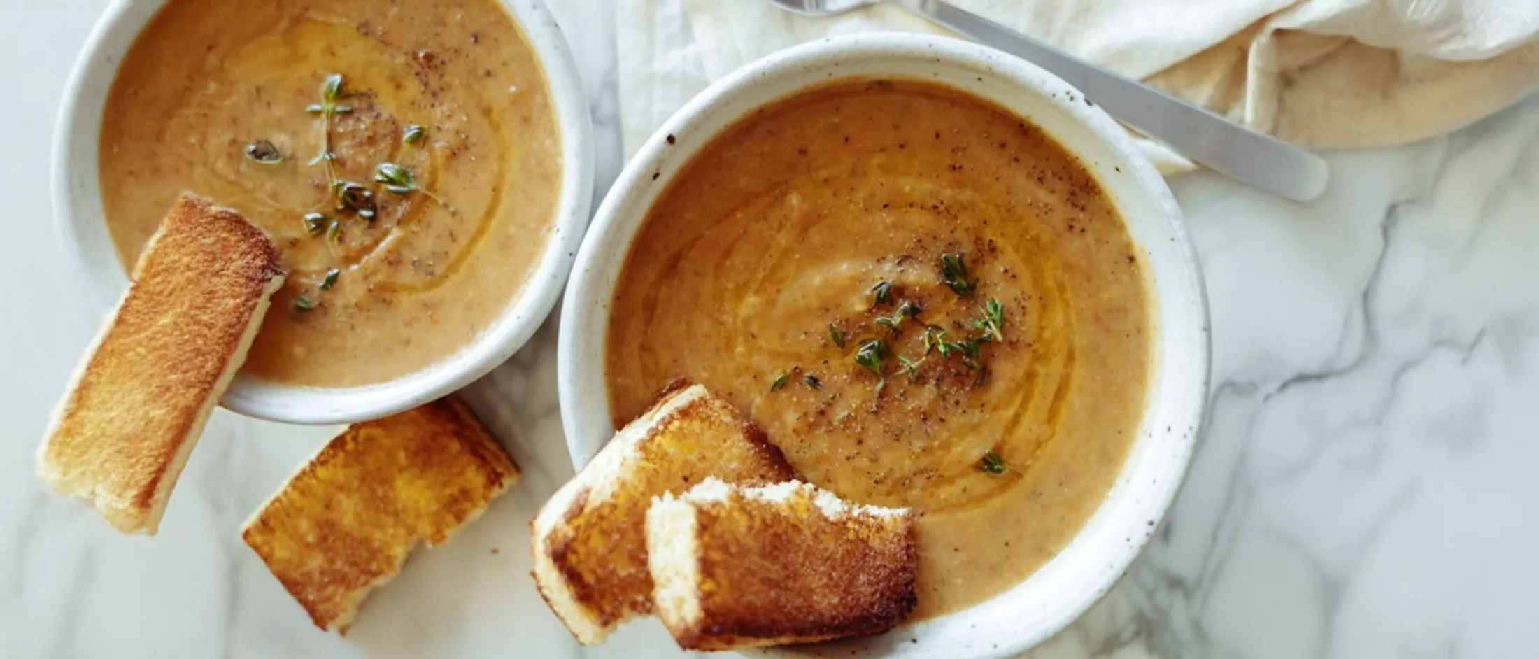 Two bowls of creamy soup garnished with herbs, served with toasted bread slices on a light marble surface.