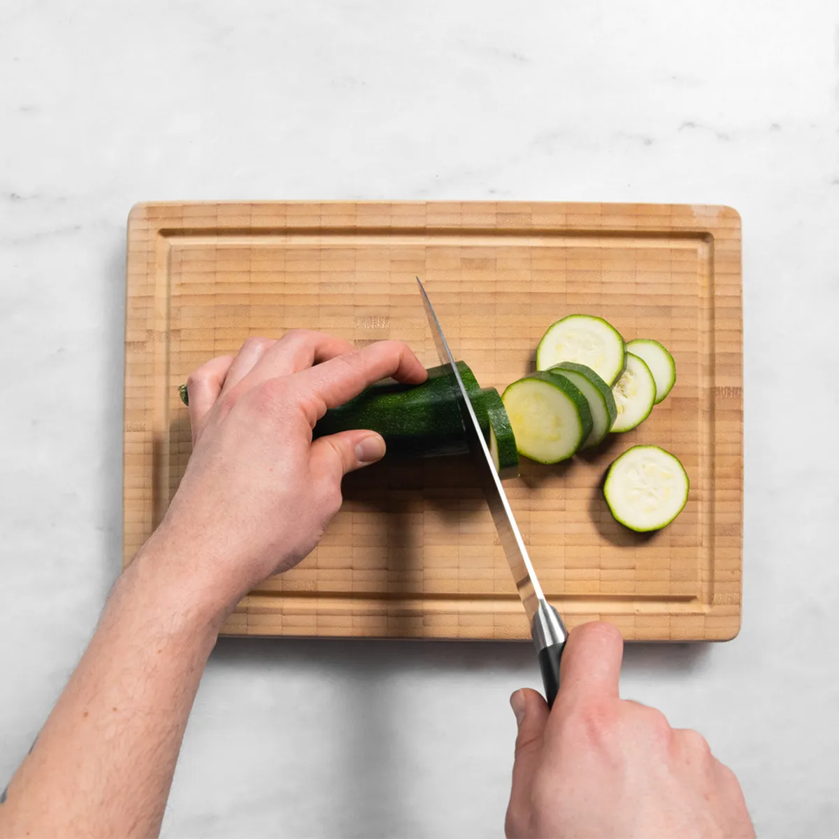Hands slicing a zucchini on a wooden cutting board with a kitchen knife.