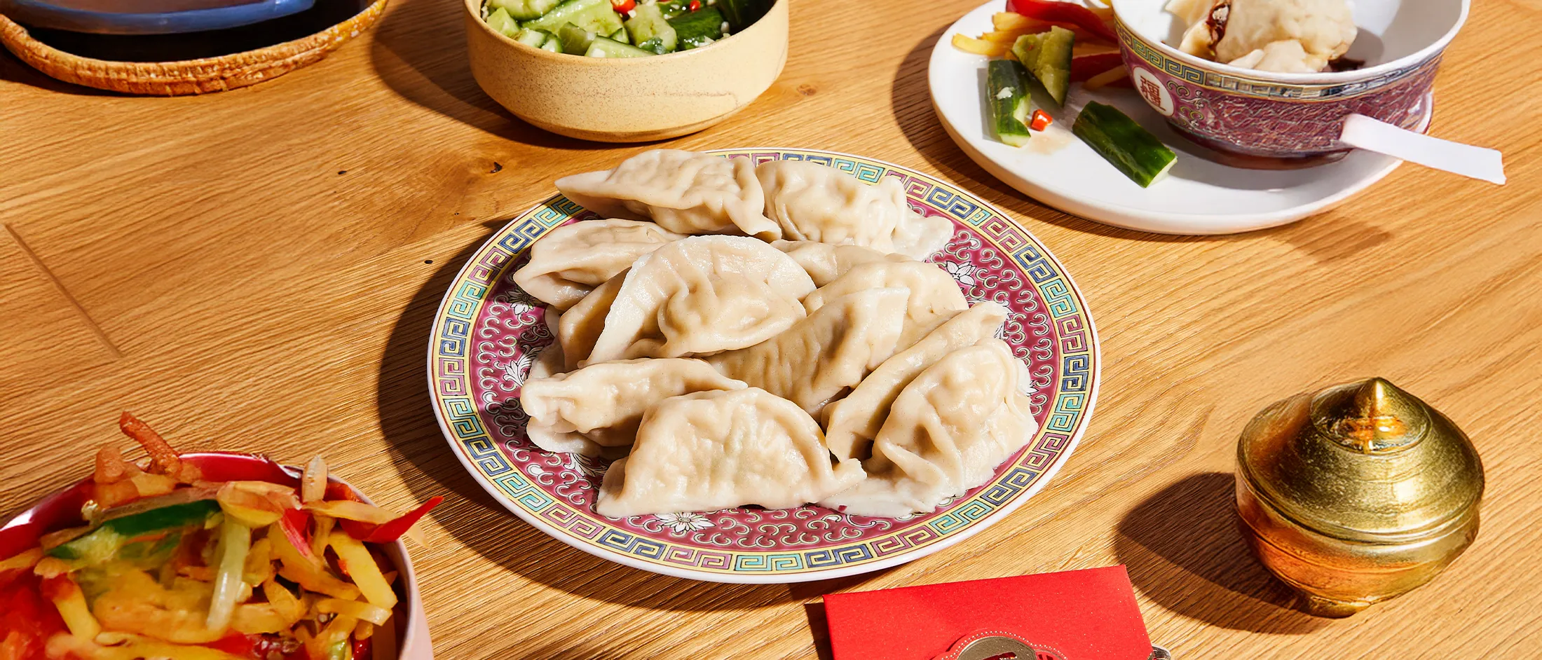 Plate of steamed dumplings on a decorative dish surrounded by small bowls of cucumber salad and mixed vegetable sides on a wooden table.