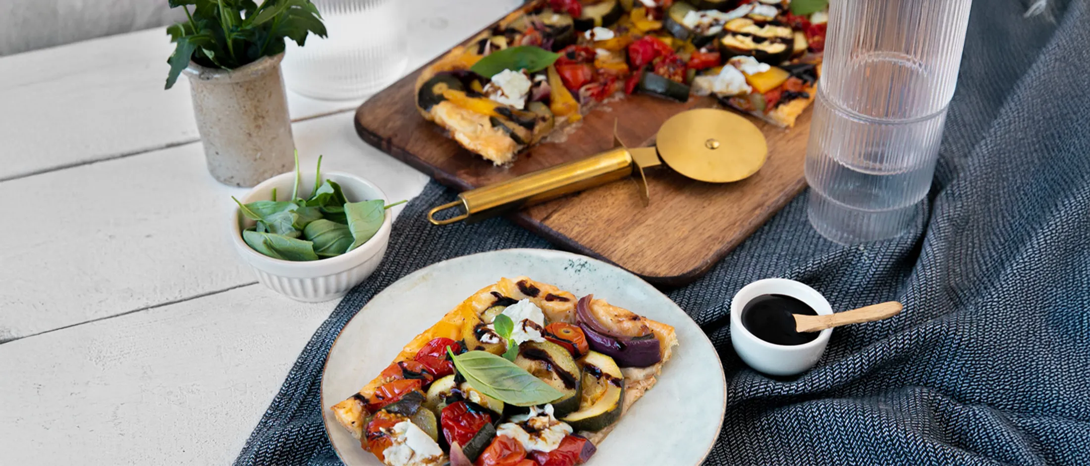 Slice of vegetable pizza with basil leaves on a plate next to a wooden pizza cutter and a bowl of dark sauce on a cloth with a wooden board holding the rest of the pizza and a small bowl of fresh basil leaves.