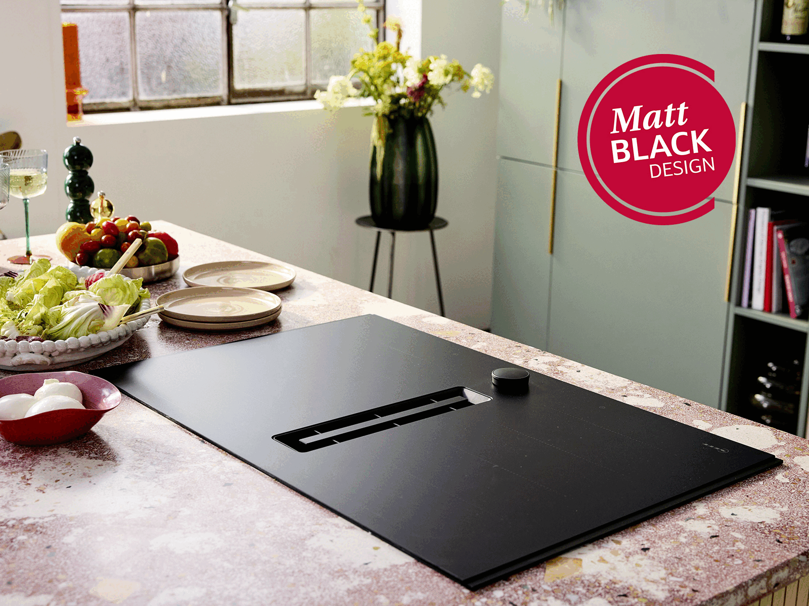 En flott, matt svart kokeplate med innebygd avtrekksvifte – en salat og tallerkener i bakgrunnen