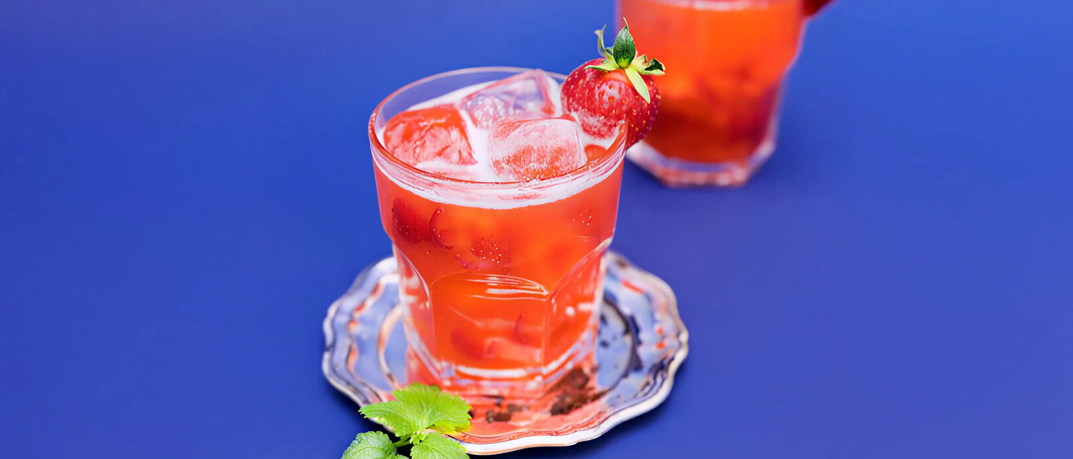 Glass of iced red fruit drink garnished with a strawberry on a silver coaster against a blue background.