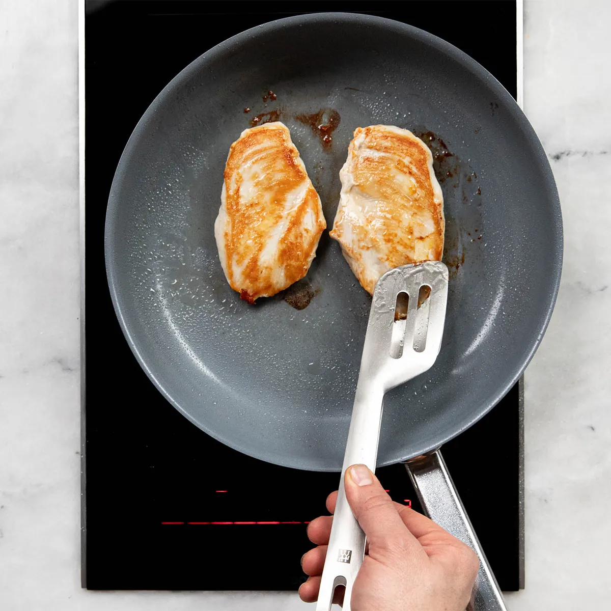 Top-down view of a hand using a metal spatula to sear two golden-brown chicken breasts in a gray nonstick frying pan set on an induction hob.